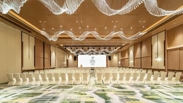 Un salón de conferencias con filas de sillas, una pantalla grande al frente y un plafón con una lámpara de diseño ondulado.