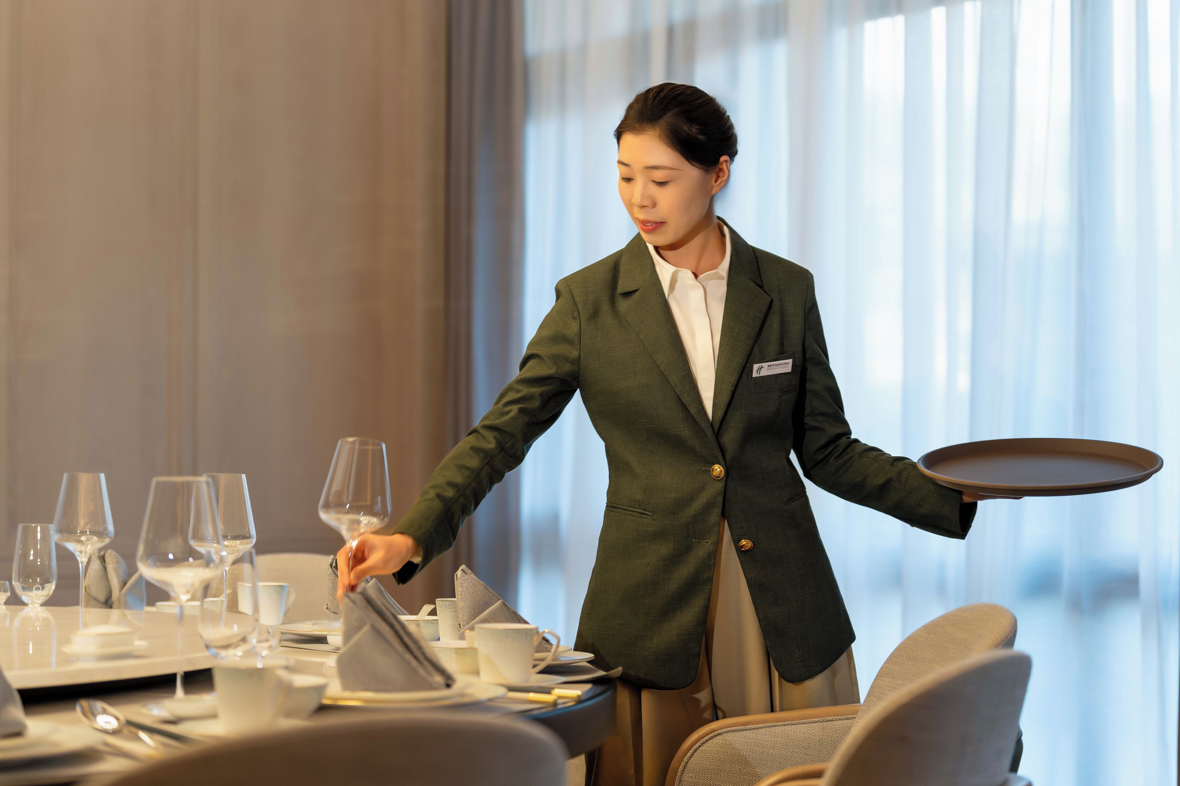 Una persona con un blazer verde y camisa blanca atendiendo una mesa con copas de vino, platos y cubiertos.