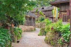 Um caminho de paralelepípedos estreito, ladeado por edifícios de madeira rústica, paredes de pedra e vegetação exuberante.