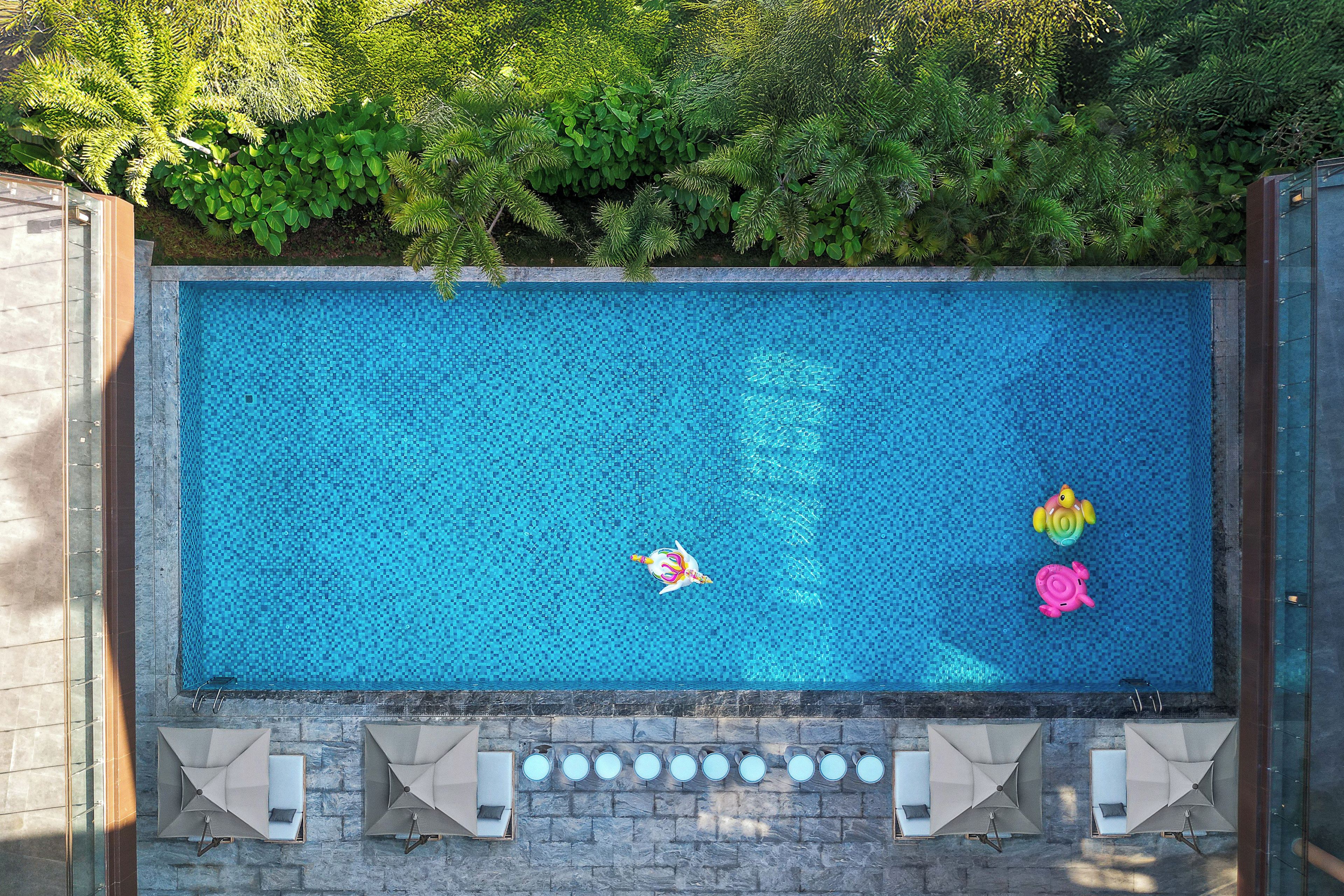 A rectangular swimming pool with a tiled surface, surrounded by a stone wall and a row of lounge chairs with umbrellas.