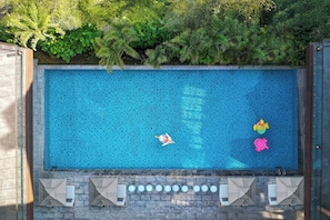 Una piscina rectangular con superficie de azulejos, rodeada por un muro de piedra y una fila de sillones de descanso con sombrillas.