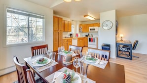 Un comedor con una mesa de madera dispuesta para una comida, una cocina al fondo y un reloj en la pared.