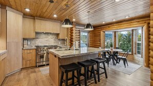 Une cuisine rustique aménagée dans une cabane en rondins, avec des meubles en bois, un îlot central doté de tabourets, une cuisinière et un coin repas comprenant une table et des chaises.