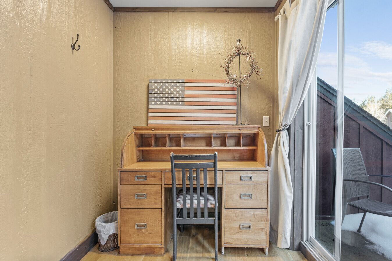 Une pièce d’angle avec un bureau en bois, une chaise, une fenêtre donnant sur un beau paysage et un drapeau américain décoratif accroché au mur.