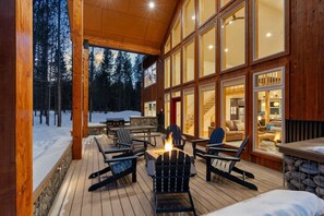 A wooden deck with outdoor seating and a fire pit, overlooking a snowy forest.
