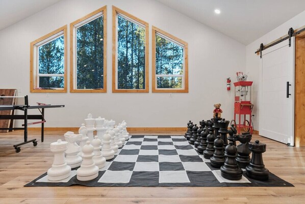 A room with a large checkered floor, a chessboard set up with white and black pieces, and three large windows with a view of trees.