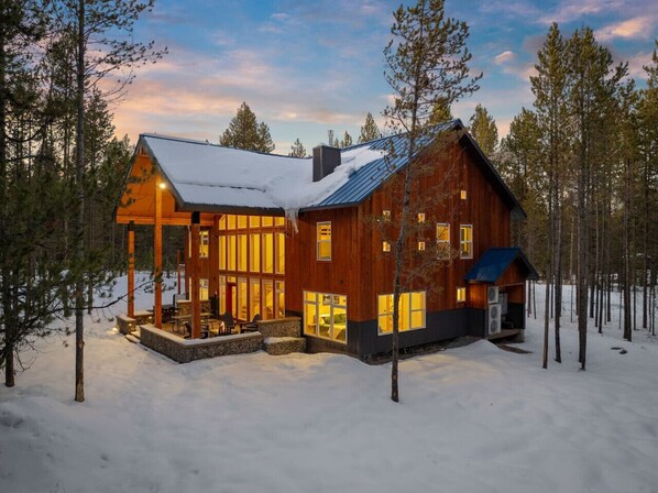 A wooden cabin with a large porch and multiple windows, surrounded by trees and snow.