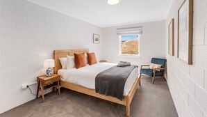Un dormitorio con cama de madera, paredes blancas, ventana con vistas, una silla y una mesita de noche con lámpara.