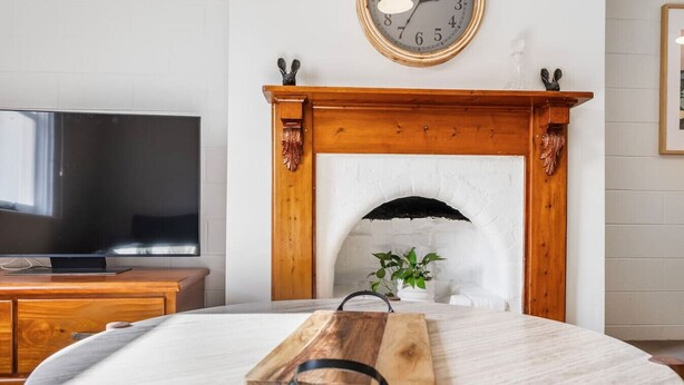 Una sala de estar con chimenea, repisa de madera, un reloj, un televisor y una tabla de cortar de madera sobre una mesa.
