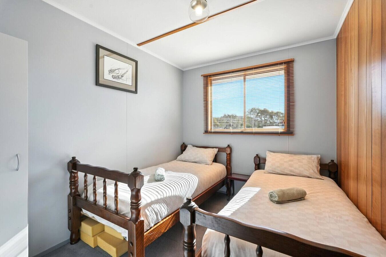 A bedroom with two wooden beds, a window with blinds, a framed picture on the wall, and a wooden headboard.