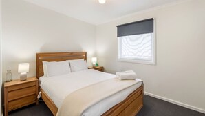 Un dormitorio ordenado con una cama de madera, dos mesitas de noche con lámparas, una ventana con persiana estampada y una pequeña mesita con un recipiente de vidrio.