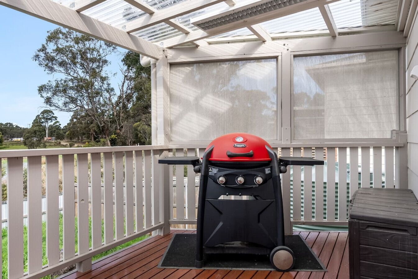 A barbecue grill on a wooden deck with a white fence and a covered area with a view of trees.