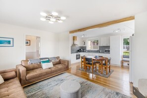 A modern living room with a brown leather sofa, a dining table with chairs, and a kitchen area with white cabinets and a stone backsplash.