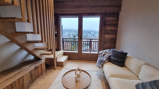 A modern living room with a staircase, a sofa, a coffee table, and a view of a cityscape through a large window. - 7 minutes from the center and superb mountain views (Megève)
