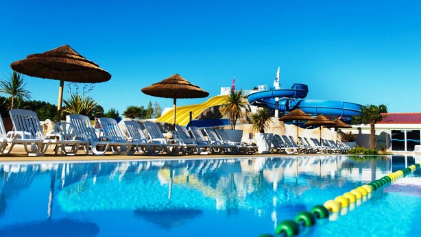 Una piscina con sillas de descanso y sombrillas, un tobogán acuático y un cielo azul despejado.