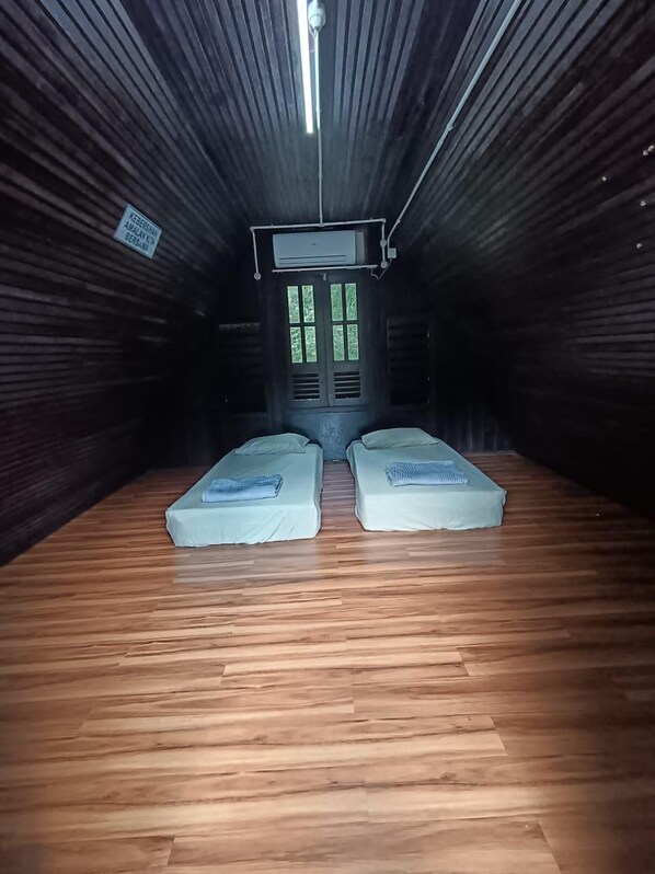 A room with two beds, a wooden floor, a window with blinds, and an air conditioning unit.