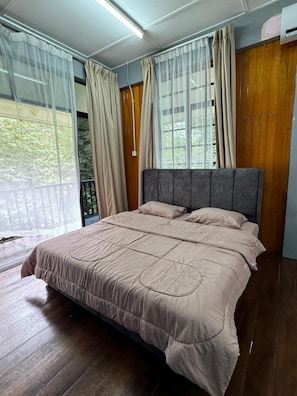 A bedroom with a large bed, wooden flooring, and a view of greenery through sheer curtains.
