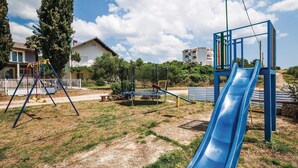 Ein Spielplatz mit einer blauen Rutsche und Schaukeln, umgeben von Gras und Bäumen, mit Gebäuden im Hintergrund.