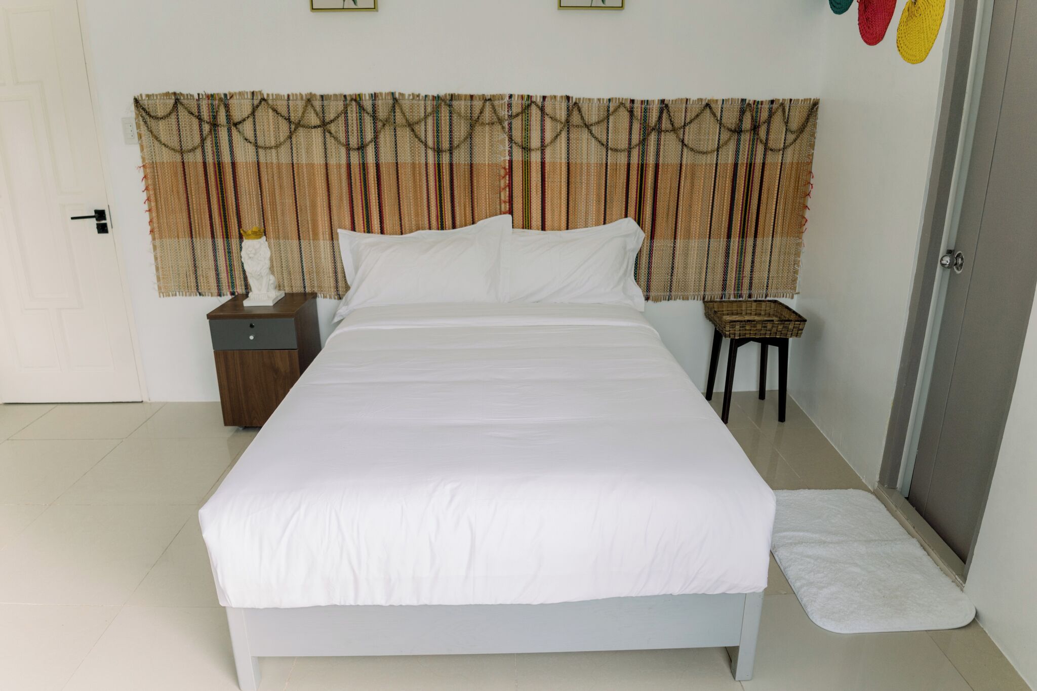 A neatly made bed with white linens, a wooden nightstand with a lamp, and a small stool with a basket beside it.
