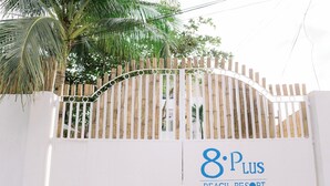 Une entrée fermée par un portail mène au complexe hôtelier 8 Plus Beach Resort. On y aperçoit une clôture blanche et, en arrière-plan, un palmier.