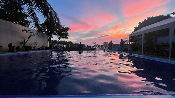 Une piscine avec vue sur le coucher de soleil, entourée de palmiers et d’un bâtiment.
