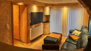 Ein gemütliches Zimmer mit Holzboden, einer Couch, einem Fernseher und einem Fenster mit Vorhängen.