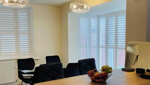 Une cuisine moderne avec un plan de travail en bois, des chaises noires et un saladier à fruits. On y trouve deux lampes suspendues et une fenêtre avec des stores.