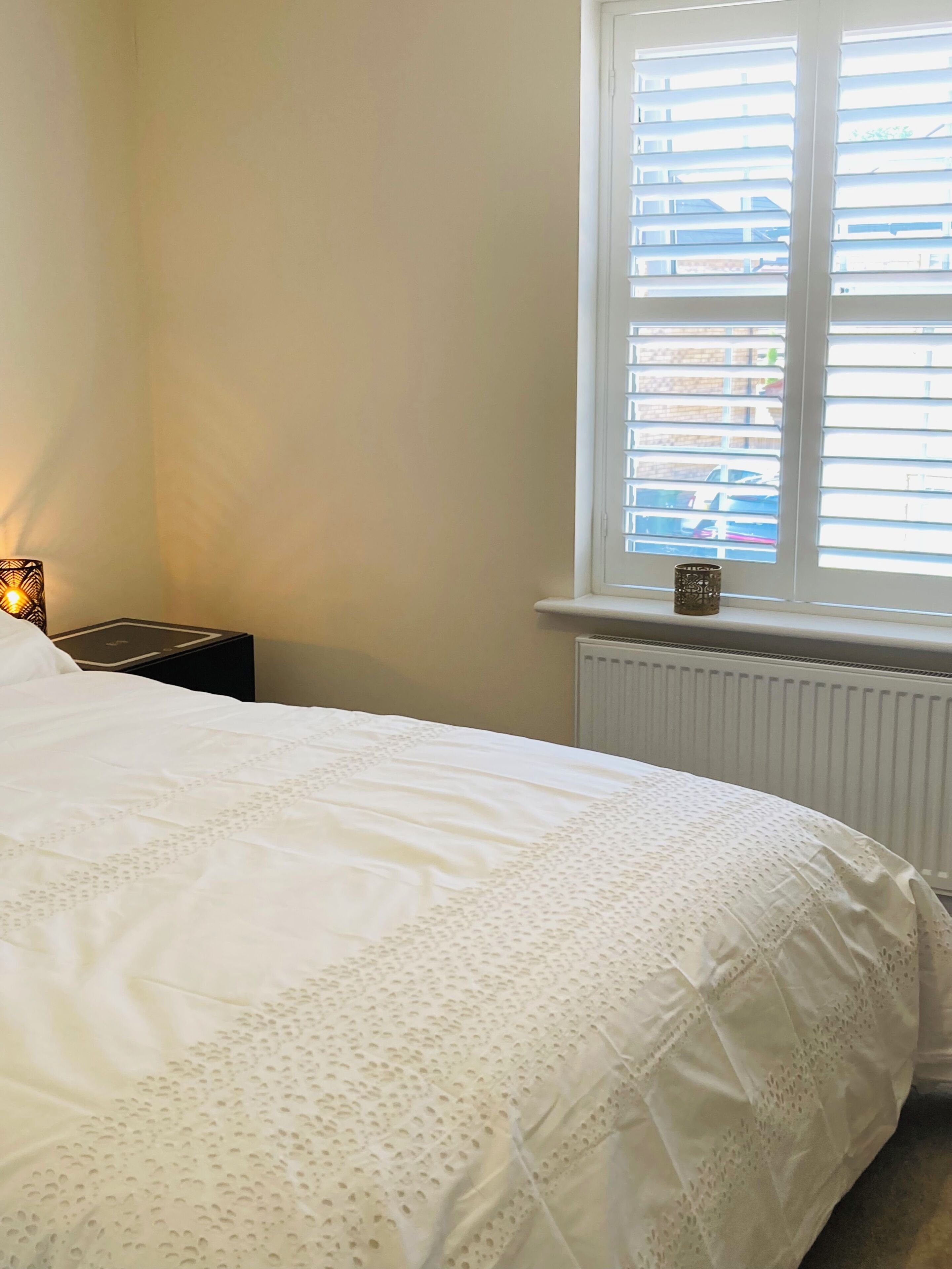 A bedroom with a bed, a nightstand, a window with blinds, and a radiator.