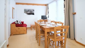 Une pièce comprenant une table à manger en bois, préparée pour quatre personnes, un canapé rouge, un téléviseur et une fenêtre avec des rideaux.