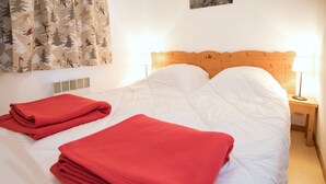 Un lit bien fait, avec du linge de lit blanc et des couvertures rouges, une tête de lit en bois, une lampe de chevet et une fenêtre avec des rideaux.