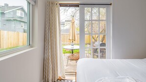 A bedroom with a large window, a sliding glass door leading to a patio with a table and chairs, and a bed with a white cover. - Charming Ballard Cottage Retreat with Cozy Fireplace (Seattle)