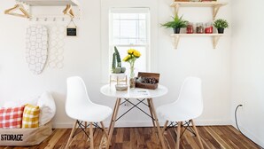A room with a round table, two white chairs, a storage bench, a window with a view, a hanging rack with clothes, and shelves with plants and jars. - Charming Ballard Cottage Retreat with Cozy Fireplace (Seattle)