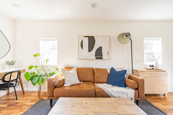 A modern living room with a brown sofa, a coffee table, and a desk with a chair. There is a large abstract painting on the wall, a floor lamp, and a window with a view of trees.
