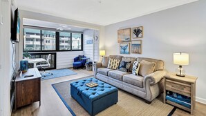 Ein Wohnzimmer mit Sofa, Hocker und Beistelltisch, ein Essbereich mit Tisch und Stühlen sowie ein großes Fenster mit Blick auf Gebäude.