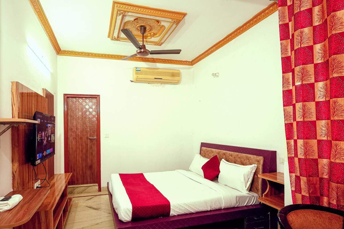 A bedroom with a bed, a TV mounted on a wooden shelf, a ceiling fan, and a door.
