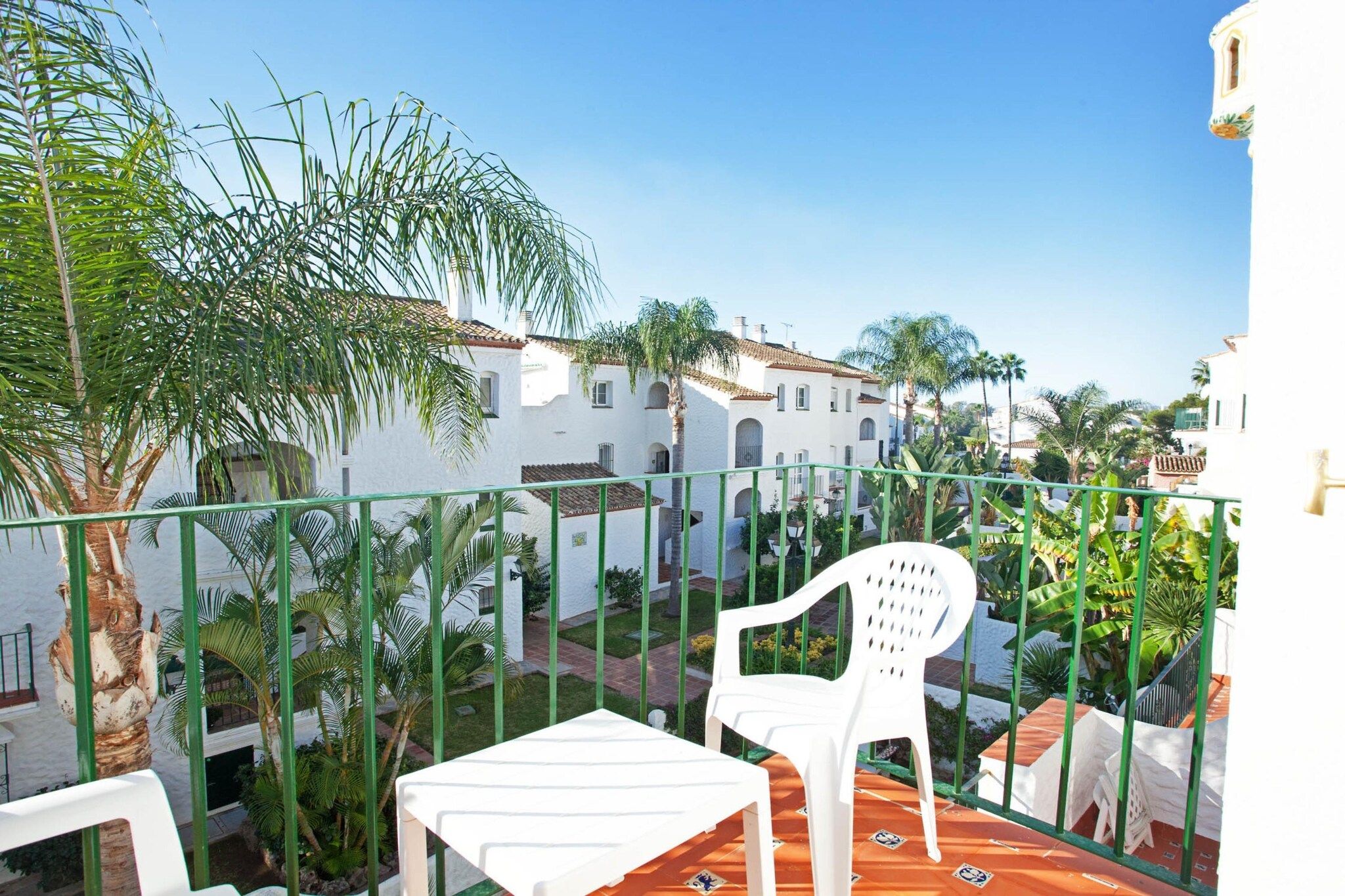 A balcony with a white plastic chair, a small table, and a view of white buildings with red-tiled roofs.