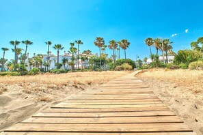 A wooden boardwalk leading to a beach with palm trees and buildings in the background. - Studio in Estepona near Sandy Beach (Estepona)