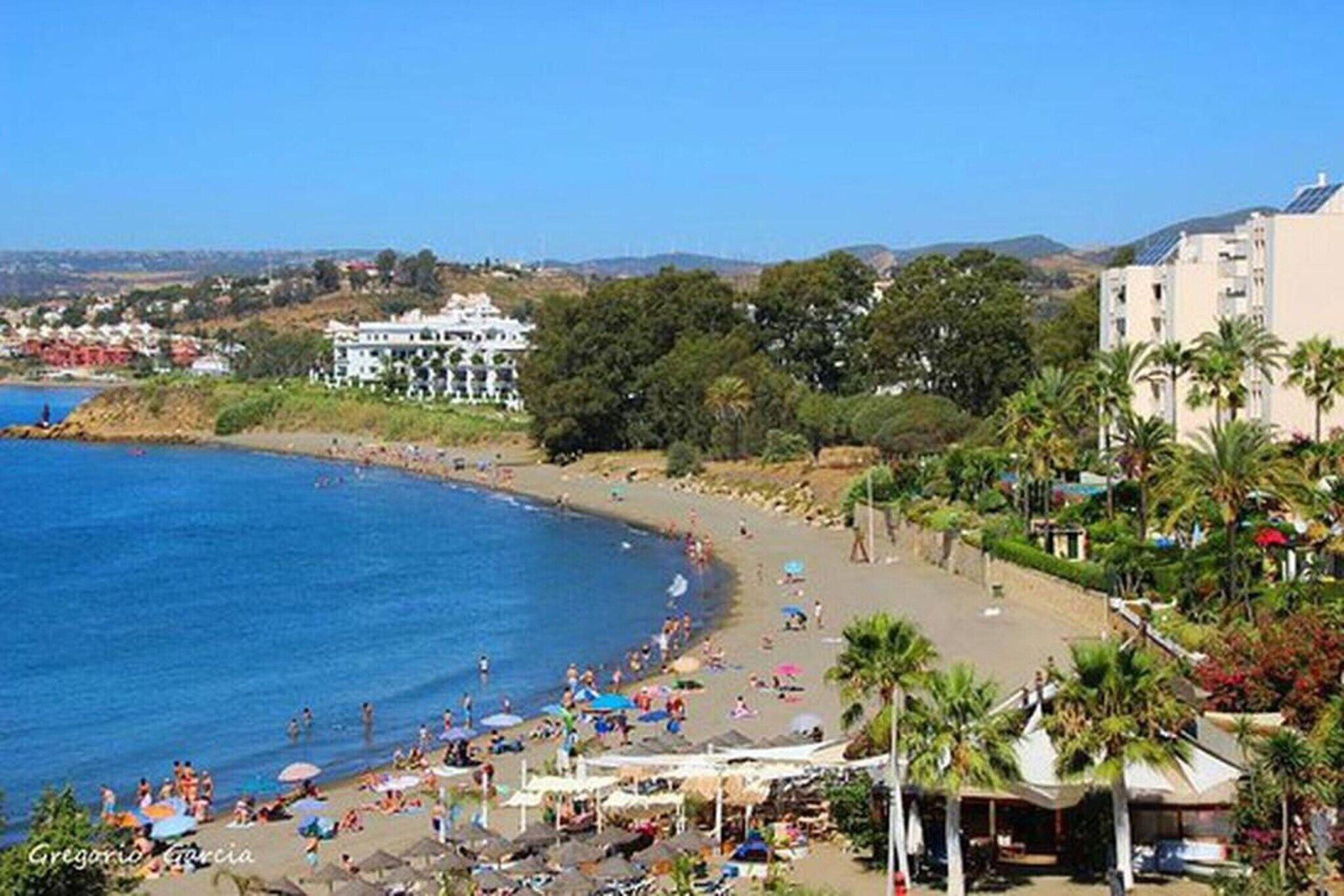 A beach with people, palm trees, and buildings in the background.