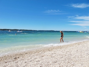 Ein Strand mit klarem türkisblauem Wasser, einem kiesigen Ufer und Booten in der Ferne.