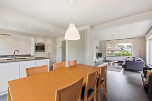 A modern kitchen with a central island, a dining area with a wooden table and chairs, and a living area with a sofa and a TV. - Villa in Koksijde Near the Beach (Koksijde)