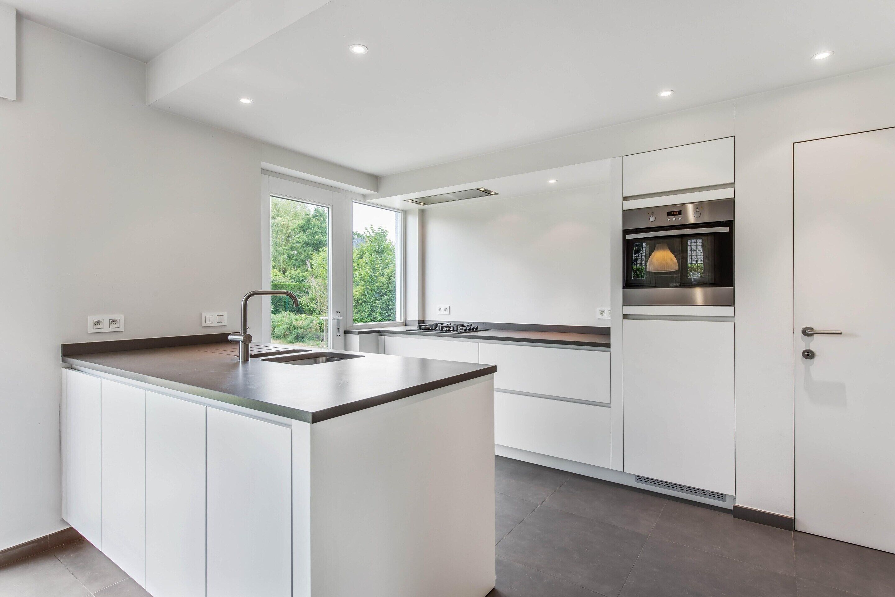 A modern kitchen with white cabinetry, a central island, a built-in oven, and a window providing natural light.