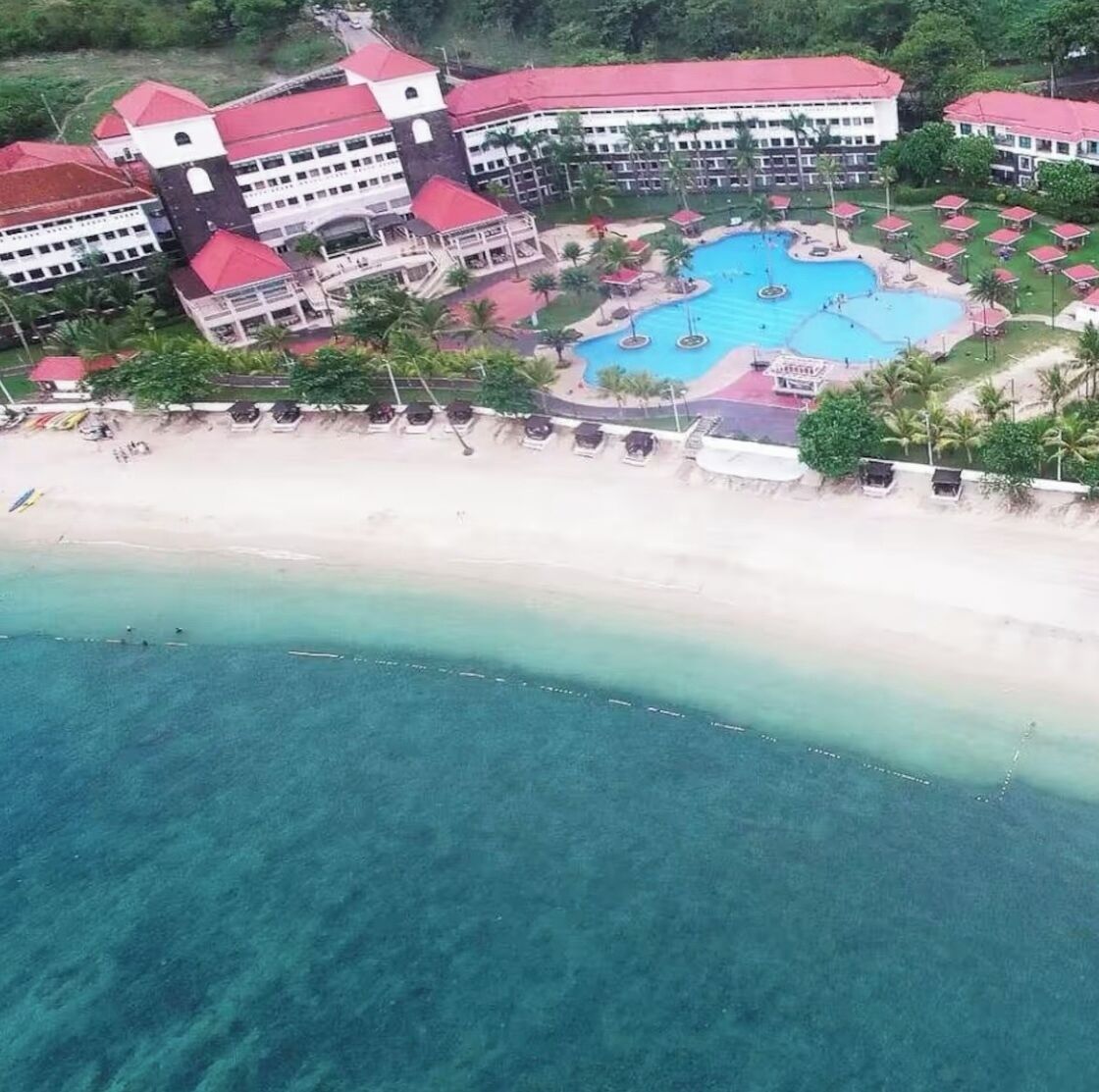 An aerial view of a resort with a large swimming pool, beachfront, and multiple buildings with red roofs.