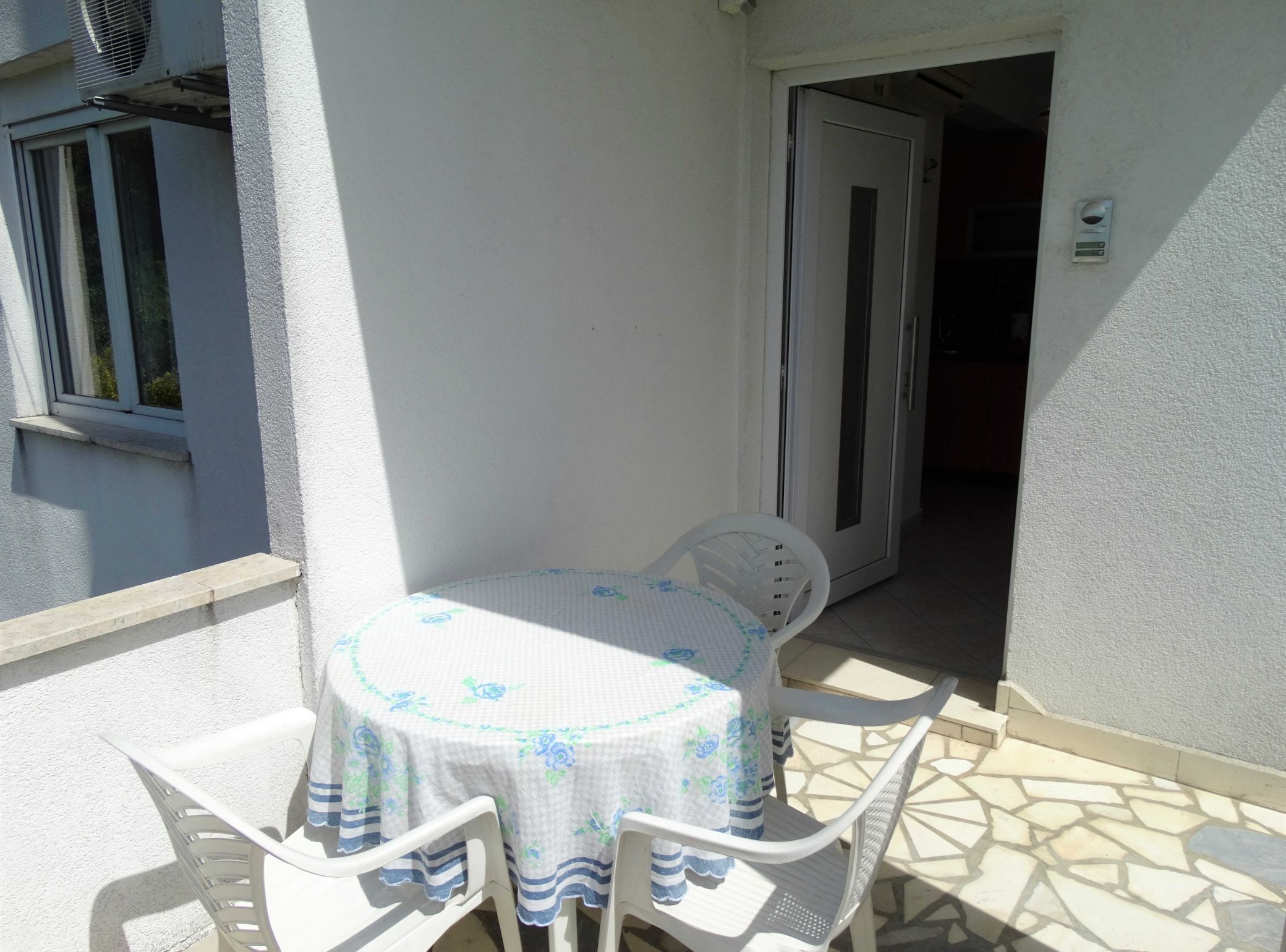 A small outdoor patio with a round table and four chairs, a door leading inside, and a window with blinds.