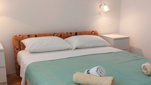 Una cama bien hecha con almohadas blancas y una colcha verde, dos toallas enrolladas sobre la cama y una mesita de noche con una lámpara.