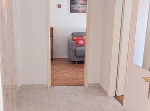 A hallway with a tiled floor leading to a room with a grey sofa and a red cushion.