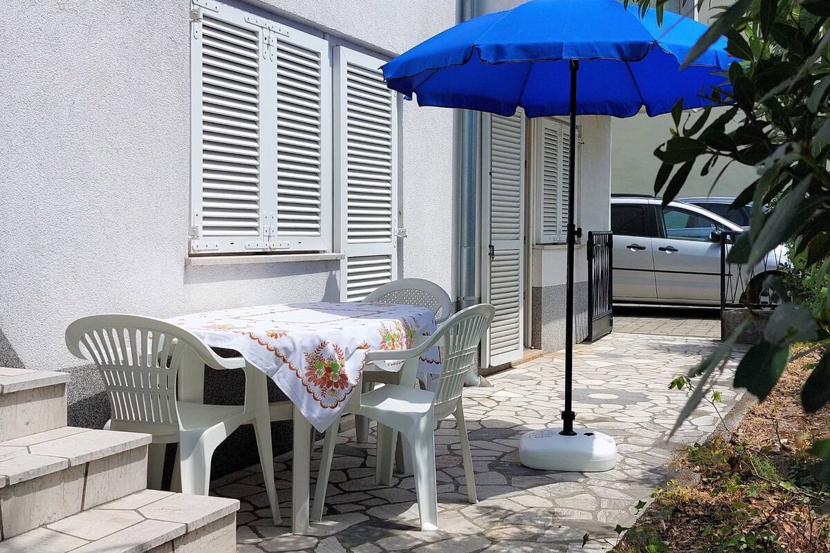 A patio with a table and chairs, a blue umbrella, and a car parked in the background.