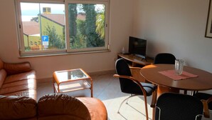 Una sala de estar con un sofá de cuero marrón, una mesa de centro de vidrio, una mesa de comedor con sillas, un televisor y vistas a edificios y árboles a través de un amplio ventanal.
