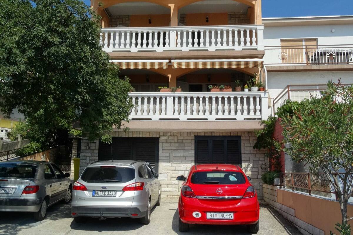 Ein zweistöckiges Gebäude mit einem Balkon im zweiten Stock. Vorne sind drei Autos geparkt.