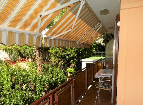Un balcon avec un auvent à rayures, une table recouverte d’une nappe à motifs floraux et une chaise.