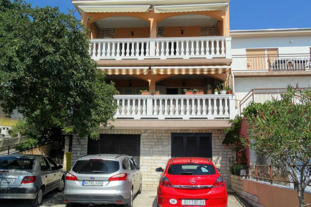 Ein zweistöckiges Gebäude mit einem Balkon im oberen Geschoss, Autos vor der Fassade und ein Baum zur Linke.